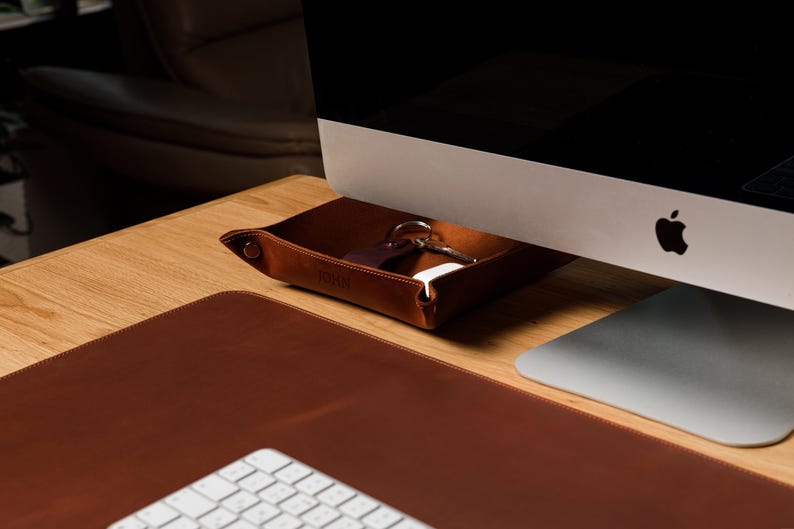 May include: A square brown leather valet tray with snap-button corners, containing a set of metal keys and a small white object. The tray is positioned on a wooden desk surface next to a computer monitor and a large brown leather desk mat.