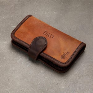 Könnte beinhalten: Ein rechteckiges Portemonnaie aus braunem Leder mit einem dunkelbraunen Lederriemen als Verschluss und einem Druckknopf. Die Vorderseite ist mit dem Wort DAD und einem kleinen kreisförmigen Blumenemblem geprägt. Die Kanten sind dunkelbraun vernäht.