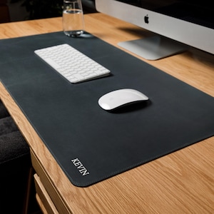 May include: A large, rectangular, dark charcoal gray desk mat with rounded corners and stitched edges. The name KEVIN is embossed in white capital letters in the bottom left corner. The mat is placed on a wooden desk surface under a computer.
