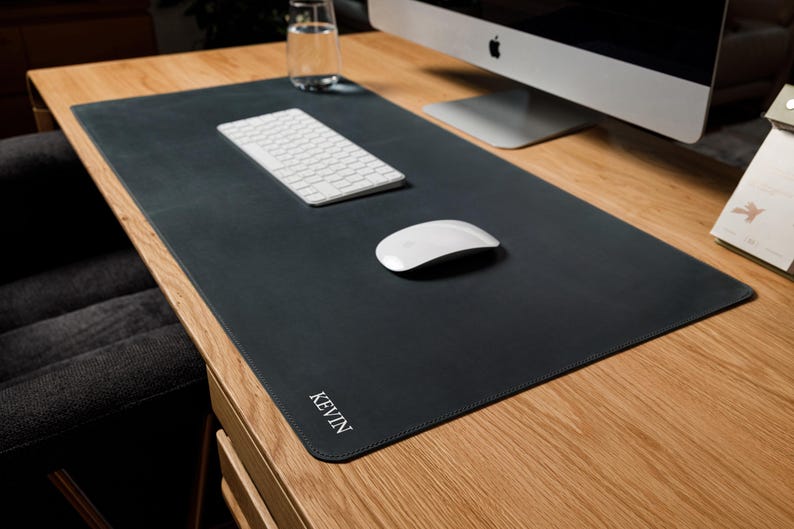 May include: A large, rectangular, dark charcoal gray desk mat with rounded corners and stitched edges. The name KEVIN is embossed in white capital letters in the bottom left corner. The mat is placed on a wooden desk surface under a computer.