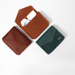 May include: Three leather cases in shades of brown and green, arranged on a white surface. One case is open, revealing white headphones. The cases are personalized with names: "JOHN", "KGP", and "LISA".