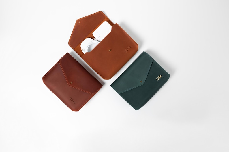 May include: Three leather cases in shades of brown and green, arranged on a white surface. One case is open, revealing white headphones. The cases are personalized with names: "JOHN", "KGP", and "LISA".