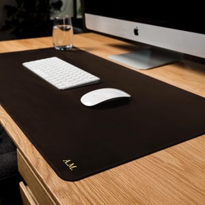 May include: A rectangular dark brown desk mat with rounded corners and stitched edges. The mat features the gold embossed initials A.M. in the bottom left corner. It is placed on a wooden desk surface under a white computer keyboard and mouse.
