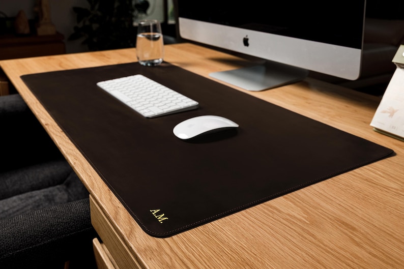 May include: A rectangular dark brown desk mat with rounded corners and stitched edges. The mat features the gold embossed initials A.M. in the bottom left corner. It is placed on a wooden desk surface under a white computer keyboard and mouse.