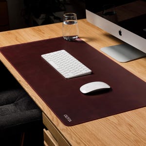 Op de afbeelding: Een rechthoekige, donkerbordeauxrode bureauonderlegger met afgeronde hoeken en de naam OLIVIA in witte letters in de linkerbenedenhoek. De mat ligt op een houten bureau en biedt een glad oppervlak voor een wit toetsenbord en een computermuis.