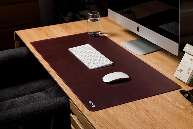 May include: A rectangular dark burgundy desk mat with rounded corners and the name OLIVIA embossed in white lettering on the bottom left corner. The mat is positioned on a wooden desk surface, providing a smooth base for a white keyboard and mouse.
