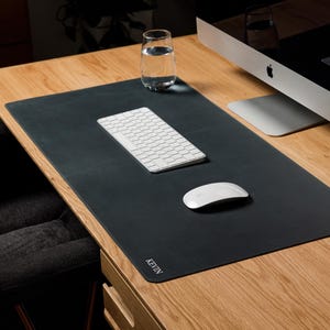 Peut inclure: Un sous-main rectangulaire noir aux coins arrondis avec le nom KEVIN imprimé en blanc dans le coin inférieur gauche. Le tapis est posé sur un bureau en bois, supportant un clavier sans fil blanc et une souris d'ordinateur sans fil blanche.