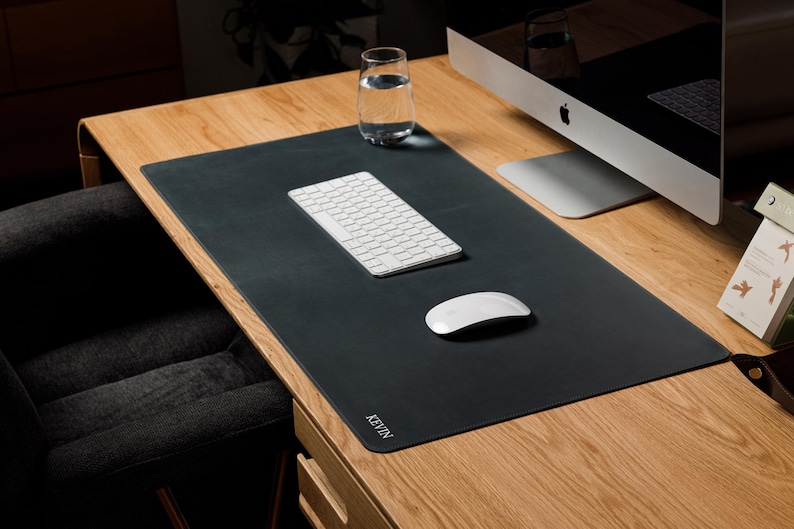 May include: A rectangular black desk mat with rounded corners and the name KEVIN printed in white in the bottom left corner. The mat is positioned on a wooden desk surface, supporting a white wireless keyboard and a white wireless computer mouse.