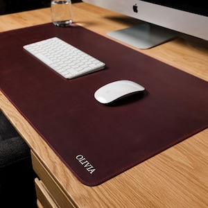 May include: A rectangular dark burgundy desk mat with rounded corners and the name OLIVIA embossed in white capital letters in the bottom left corner. The mat features a smooth, solid-colored surface and is positioned on a light wood desk.
