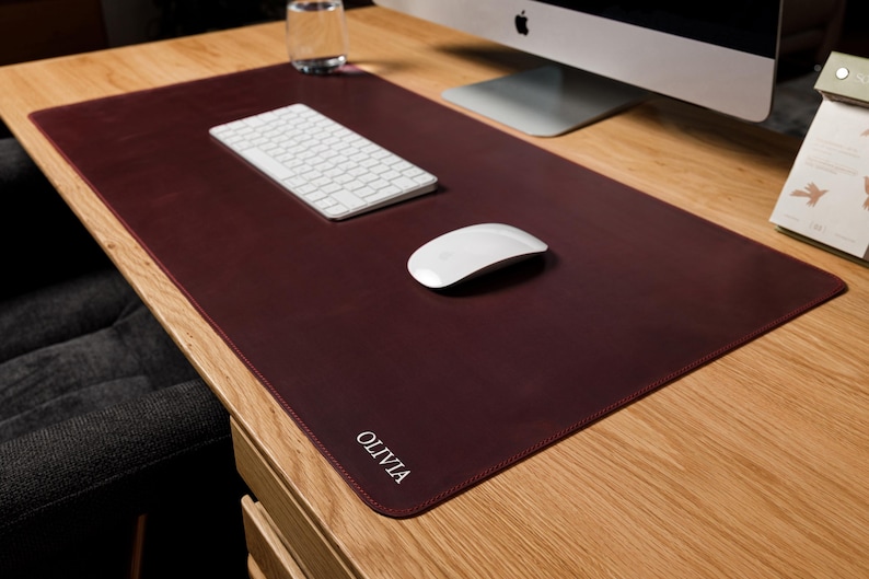 May include: A rectangular dark burgundy desk mat with rounded corners and the name OLIVIA embossed in white capital letters in the bottom left corner. The mat features a smooth, solid-colored surface and is positioned on a light wood desk.