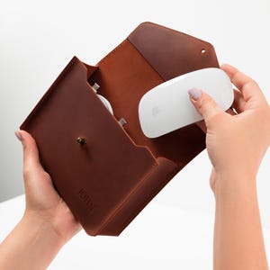 May include: A brown leather envelope-style case featuring a brass stud closure and the embossed name JOHN. The case contains a white computer mouse and a white charging cable, showcasing a structured design with visible stitching along the edges.