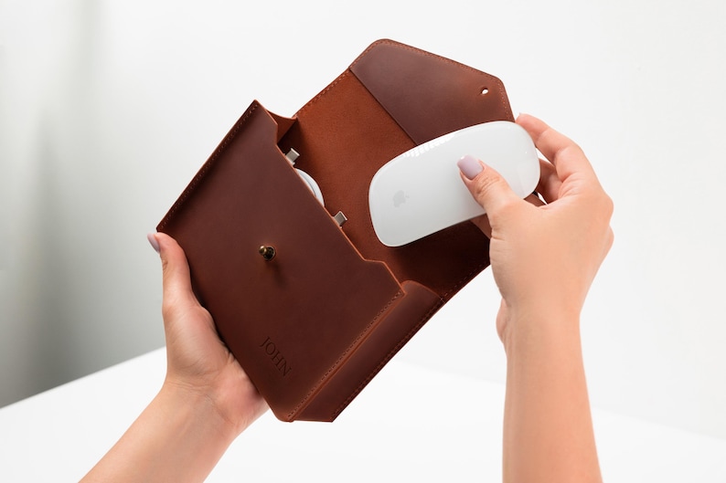 May include: A brown leather envelope-style case featuring a brass stud closure and the embossed name JOHN. The case contains a white computer mouse and a white charging cable, showcasing a structured design with visible stitching along the edges.
