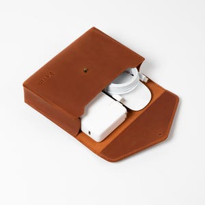 May include: A brown leather tech organizer with a snap closure, open to reveal a white charger, mouse, and cable. The word "Kevin" is embossed on the front. The organizer is on a white background.