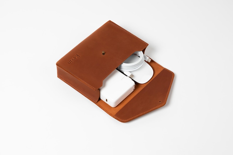 May include: A brown leather tech organizer with a snap closure, open to reveal a white charger, mouse, and cable. The word "Kevin" is embossed on the front. The organizer is on a white background.