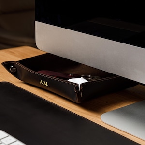 May include: A dark brown leather valet tray with snap-button corners and the gold-embossed initials A.M. on the front edge. The tray is positioned under a computer monitor and contains small personal items like keys and a white charging case.
