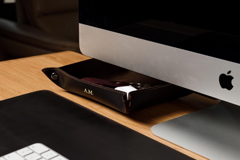 May include: A dark brown leather valet tray with snap-button corners and the gold-embossed initials A.M. on the front edge. The tray is positioned under a computer monitor and contains small personal items like keys and a white charging case.