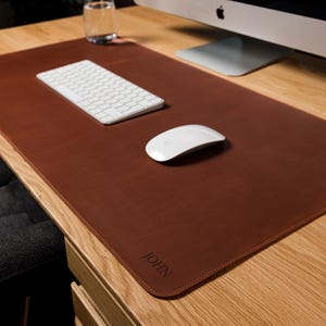 May include: A rectangular brown leather desk mat with rounded corners and stitched edges. The mat features the name JOHN embossed in the bottom right corner. It is placed on a wooden desk surface with a white keyboard and a white computer mouse.