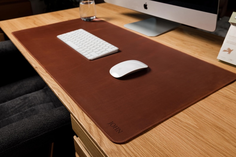 May include: A rectangular brown leather desk mat with rounded corners and stitched edges. The mat features the name JOHN embossed in the bottom right corner. It is placed on a wooden desk surface with a white keyboard and a white computer mouse.