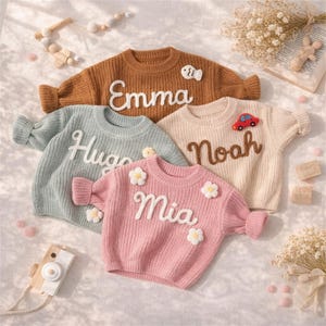 May include: Four children's sweaters in shades of brown, blue, pink, and cream. Each sweater has a name embroidered on the front in white script. The sweaters are decorated with small appliques, including a fish, a car, and flowers.