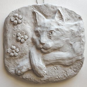 May include: A light gray clay wall plaque featuring a sculpted cat resting among embossed flowers. The piece has a metal hanging loop at the top. A circular stamp in the corner reads: ORIGINAL DESIGN 100% Handmade by the Artist *ONLY ON WIREANIMALS.