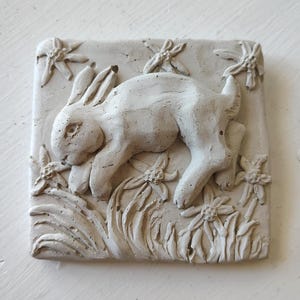Puede incluir: Escultura en relieve cuadrada de color blanquecino con un conejo entre flores y follaje estilizados. El conejo está esculpido en posición reclinada. La escultura es texturizada y tiene un aspecto hecho a mano. El texto "ORIGINAL DESIGN 100% Handmade by the Artist ONLY on WIREANIMALS" está en un círculo.