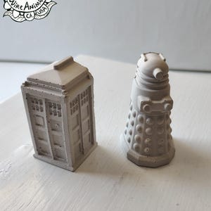 May include: Two small, light grey concrete sculptures. One is a TARDIS, a British police box, and the other is a Dalek, a fictional extraterrestrial race. The sculptures are handmade by Wire Animals, CA, USA.