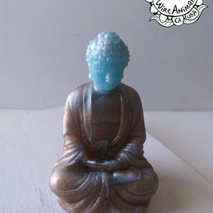 May include: A small, blue and gold, hand-crafted, wire sculpture of a meditating Buddha. The sculpture is sitting on a white surface. The sculpture is labeled "100% Handmade by Wire Animals CA. USA."
