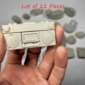 May include: A small, off-white miniature boombox with speaker details and a cassette deck is held in a hand. The text "Lot of 22 Pieces" is written in red at the top. Other small, gray pieces are in the background.