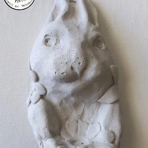 May include: White ceramic sculpture of a rabbit's head with a floral design around the neck. The sculpture is handmade and has a textured surface. The text "ORIGINAL DESIGN 100% HANDMADE by the ARTIST ONLY ON WIREANIMALS" is visible in a circle around the top of the sculpture.