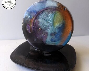 May include: A round, black resin coaster with a colourful, abstract design. The design features swirls of blue, green, purple, and gold. The coaster is on a black stand.
