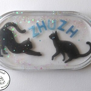 May include: A clear oval resin coaster featuring two black cat silhouettes and blue glitter. The word "ZHUZH" is spelled out in blue letters. The coaster has a white background with iridescent glitter.