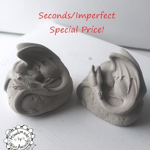 May include: Two gray concrete dragon sculptures, each with a curled tail and wings, resting on a rock-like base. The sculptures are handmade by Wire Animals CA USA. The text "Seconds/Imperfect Special Price!" is displayed above the sculptures.