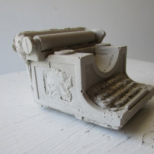 Puede incluir: Una escultura de hormig&oacute;n blanco de una m&aacute;quina de escribir vintage. La escultura tiene un dise&ntilde;o detallado con un rodillo y teclas visibles.