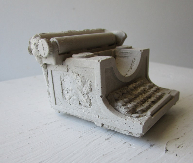 Puede incluir: Una escultura de hormig&oacute;n blanco de una m&aacute;quina de escribir vintage. La escultura tiene un dise&ntilde;o detallado con un rodillo y teclas visibles.
