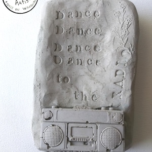 May include: A gray concrete sculpture of a boombox with the words "Dance Dance Dance Dance to the RADI" written on it.
