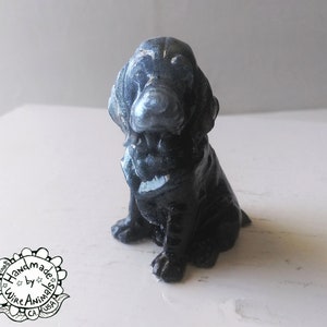 May include: A small, black, ceramic dog figurine with a sitting pose. The dog has a short, stubby tail and a slightly tilted head. The figurine is painted with a dark gray glaze and has a slightly textured surface.