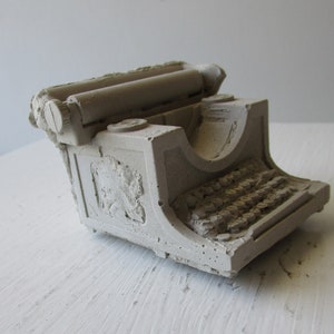 Puede incluir: Una peque&ntilde;a escultura de hormig&oacute;n gris de una m&aacute;quina de escribir vintage. La m&aacute;quina de escribir tiene una forma redondeada y un dise&ntilde;o detallado, que incluye un rodillo, teclas y un elemento decorativo en el lateral.