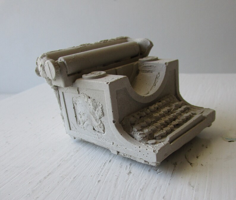 Puede incluir: Una peque&ntilde;a escultura de hormig&oacute;n gris de una m&aacute;quina de escribir vintage. La m&aacute;quina de escribir tiene una forma redondeada y un dise&ntilde;o detallado, que incluye un rodillo, teclas y un elemento decorativo en el lateral.
