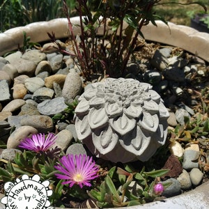 May include: A gray concrete flower sculpture with a detailed design, sitting in a pot with rocks and small pink flowers. The sculpture is handmade by Wire Animals, CA, USA.