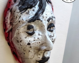 May include: A sculpted face mask with a silver metal loop for hanging. The mask features a white base with black speckles, black hair, and red accents. The text "ORIGINAL DESIGN 100% Handmade by the ARTIST" is in a circle.