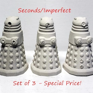 May include: Three grey concrete Dalek sculptures. Each has a domed head, a central body with circular details, and a rectangular base. The text "Seconds/Imperfect" and "Set of 3 - Special Price!" are visible.