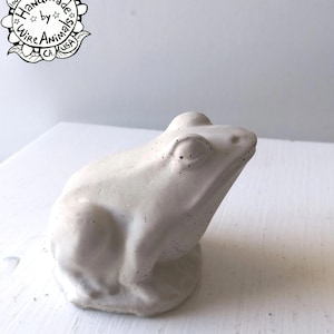 May include: A white concrete frog figurine with a textured surface. The frog is sitting on a white surface.