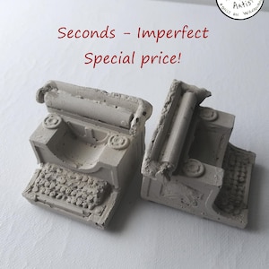 May include: Two small, light grey concrete typewriter sculptures. The typewriters have detailed keys and circular side details. Red text reads "Seconds - Imperfect Special price!" and a circular logo.