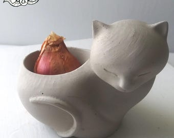 May include: A light grey, matte-finish ceramic planter shaped like a sleeping cat with a hollow body. A single brown shallot is placed inside the opening of the planter. A circular logo in the top left corner reads 100% Handmade by Wire Animals CA USA.