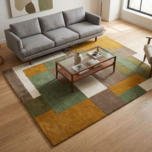 May include: A living room scene with a grey sofa, a wooden coffee table, and a multi-coloured geometric rug. The rug's design incorporates rectangular blocks in green, gold, brown, and white. The coffee table has a glass top and is made of wood.