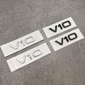 May include: Four V10 emblem stickers. Two are silver, and two are black. Each sticker has the letters "V10" in a bold, sans-serif font. The stickers are on white backing, arranged on a gray surface.