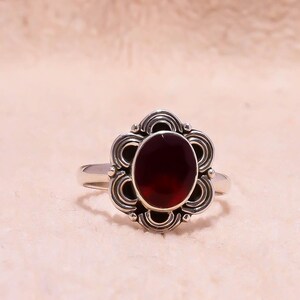Puede incluir: Un anillo de plata con una piedra preciosa ovalada de color rojo intenso. La piedra preciosa está engastada en un engaste decorativo en forma de flor con intrincados adornos. La banda del anillo es de plata pulida.