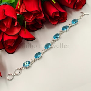 May include: A silver bracelet featuring six oval-cut, light blue gemstones set in metal bezels, connected by small circular links and a toggle clasp. The text Shibom Jeweller is visible in the center. The bracelet is displayed near red roses.