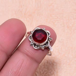 May include: A silver ring featuring a large, round, deep red gemstone set in a decorative floral-inspired setting. The ring is held between fingers, showcasing the intricate details of the metalwork and the vibrant color of the stone.