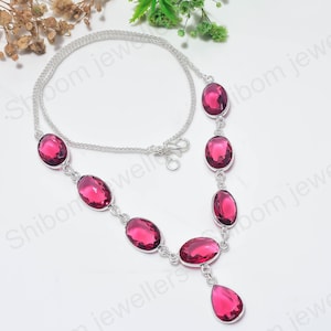 Pode incluir: Um colar de corrente prateada com sete pedras preciosas rosa-escuras, facetadas e de formato oval, dispostas na parte frontal, com um único pingente em forma de gota, facetado e rosa-escuro, pendurado no centro do design.